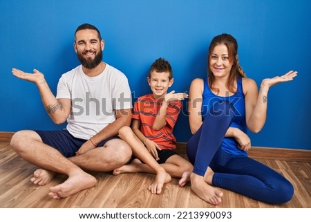 Family of three sitting on the floor at home smiling cheerful presenting and pointing with palm of hand looking at the camera. 