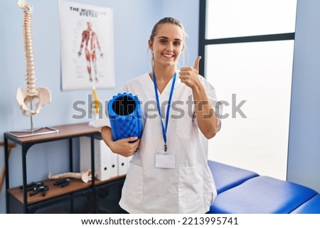 Young physiotherapist woman holding foam roll at the clinic smiling happy and positive, thumb up doing excellent and approval sign 