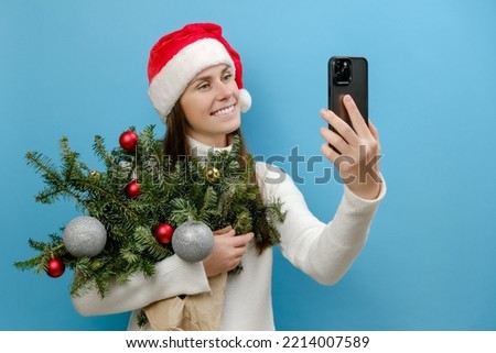 Portrait of positive cute young brunette woman in Santa Claus hat holding bouquet of spruce branches do selfie shot on mobile cell phone post photo on social network, posing on blue studio background