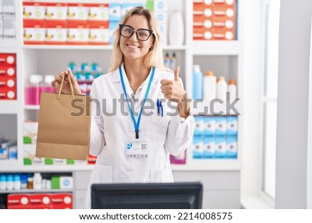 Young blonde woman working at pharmacy drugstore holding paper bag smiling happy and positive, thumb up doing excellent and approval sign  Royalty-Free Stock Photo #2214008575