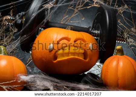 Carved Halloween pumpkin lifting a heavy barbell dumbbell. Weightlifting carving idea with exhausted effort face cut out. Gym weight workout and sport training concept. Healthy lifestyle composition.