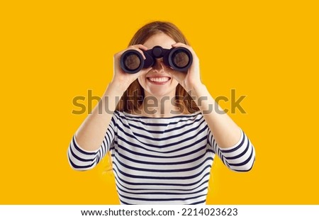 Studio shot of happy cheerful beautiful young woman in casual striped top standing isolated on yellow color background, holding modern binoculars, looking in distance and smiling Royalty-Free Stock Photo #2214023623