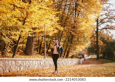Happy young woman photographing tree with yellow leaves with a smart phone while walking in park during sunny weather in autumn