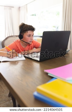 Vertical picture of caucasian boy learning and using laptop and headphones in living room. Home educations, distance educations.