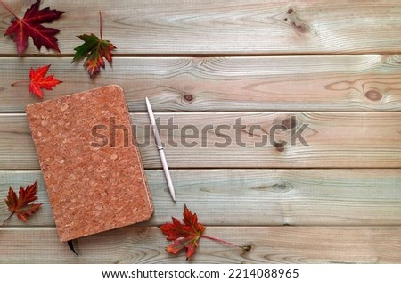 Cork notepad and autumn maple leaves. Layout. Copy space for text. Closed notebook and a pen lie surrounded by red leaves on a wooden background. Top view. Concept of autumn creativity and mood.