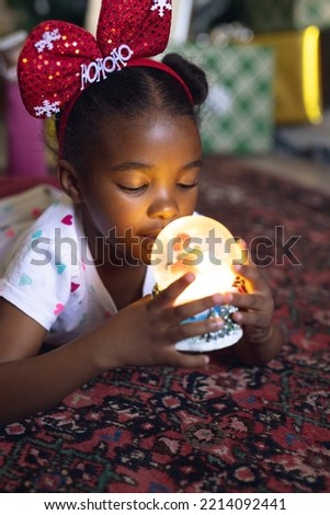 Vertical picture of african american girl holding christmas snowing ball and siting next fireplace. Christmas, childhood and celebration concept.
