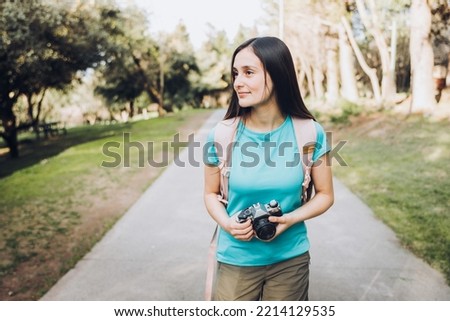 Young tourist woman traveler, wearing turquoise t shirt and a backpack, taking photos while walking in the park.