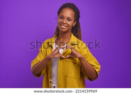 Young pretty African American woman student makes heart shape from fingers showing love and wishes you good and well-being or romantic mood dressed in casual style stands on purple background