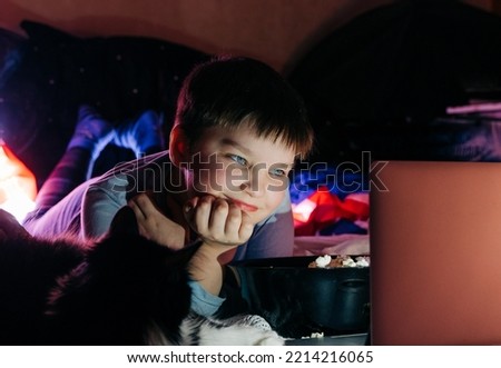 Cheerful teen boy using laptop at home watching funny movie cartoon eat pop corn, browsing internet on sofa in living room. Smiling kid looking at screen having fun enjoying weekend night vacation.