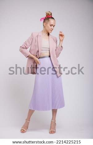 High fashion photo of a beautiful elegant young woman in pretty pink jacket, blazer, lilac lavender skirt posing on white, soft gray background. Studio Shot. Slim Figure, Blonde