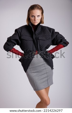 High fashion photo of a beautiful elegant young woman in a pretty white skirt with black pattern, jacket, red sweater posing on soft gray background. Studio Shot. Redhead, freckles