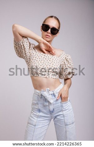 High fashion photo of a beautiful elegant young woman in pretty blue denim jeans, beige top with floral pattern, stylish sunglasses, gathered hair on white, soft gray background. Slim figure. Blonde.