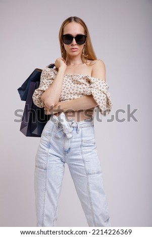 High fashion photo of a beautiful elegant young woman in pretty blue denim jeans, beige top with floral pattern, stylish sunglasses on white, soft gray background. Slim figure. Blonde. Shopping bags