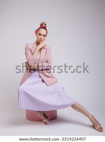 High fashion photo of a beautiful elegant young woman in pretty pink jacket, blazer, lilac lavender skirt posing on white, soft gray background. Slim Figure, Blonde. Model sits on cylinder, cube