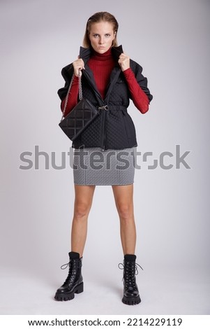 High fashion photo of a beautiful elegant young woman in a pretty white skirt with black pattern, jacket, red sweater, handbag on soft gray background. Studio Shot. Redhead, freckles