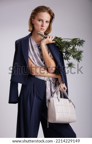 High fashion photo of a beautiful elegant young woman in a pretty blue suit, jacket, blazer, trousers, pants, handbag, scarf with a pattern, flowers bouquet posing over white, soft gray background. 