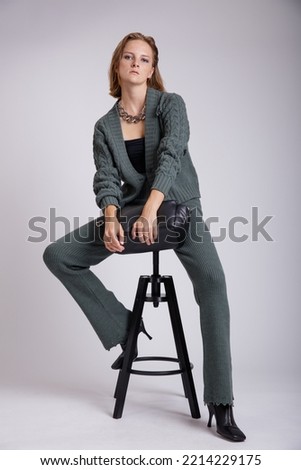 High fashion photo of a beautiful elegant young woman in a pretty green knitwear sweater, pants, trousers, leather handbag, black top posing over white, soft gray background. Studio Shot, portrait. 