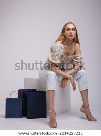 High fashion photo of a beautiful elegant young woman in pretty blue denim jeans, beige top with floral pattern posing on white, soft gray background. Blonde. Model sits on cube. Shopping bags