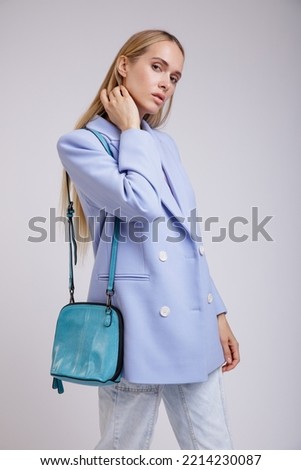 High fashion photo of a beautiful elegant young woman in a pretty jacket, blazer, denim jeans, aqua blue handbag posing over white, soft gray background. Studio Shot.	