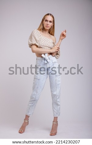 High fashion photo of a beautiful elegant young woman in pretty blue denim jeans, beige top with floral pattern posing on white, soft gray background. Slim Figure, Blonde.	
