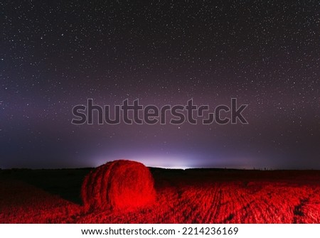 Glowing Stars And Sunset Lights Hay Bales After Harvest. Agricultural Colorful Background Copy Space. Night Starry Sky Above Rural Landscape Field Meadow With Rolls Of Straw In Fields After Harvest.
