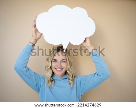 beautiful young woman with curly blond hair stands in front of brown wall and holds white speech bubble, thought bubble