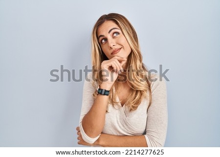 Young blonde woman standing over isolated background with hand on chin thinking about question, pensive expression. smiling with thoughtful face. doubt concept. 