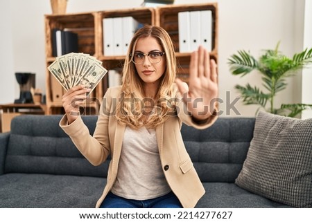 Young blonde therapist woman working at therapy office holding money with open hand doing stop sign with serious and confident expression, defense gesture 