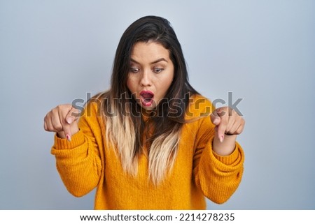 Young hispanic woman standing over isolated background pointing down with fingers showing advertisement, surprised face and open mouth 