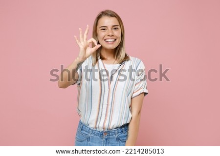 Funny young blonde woman girl in casual striped shirt posing isolated on pastel pink wall background studio portrait. People sincere emotions lifestyle concept. Mock up copy space. Showing OK gesture