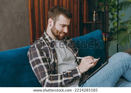 Young happy graphic designer man he wears shirt hold in hand work use write draw stylus pc pen on tablet pc computer sit on blue sofa in own living room apartment stay home indoor flat on weekends.