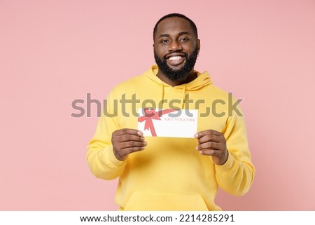 Smiling cheerful young african american man 20s wearing casual yellow streetwear hoodie standing hold in hand gift certificate looking camera isolated on pastel pink color background studio portrait