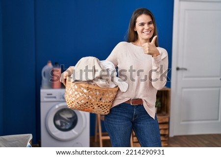 Beautiful woman holding laundry basket smiling happy and positive, thumb up doing excellent and approval sign 