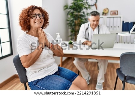 Senior woman sitting at doctor appointment praying with hands together asking for forgiveness smiling confident. 