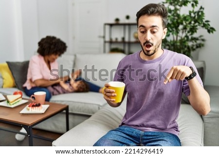 Hispanic father of interracial family drinking a cup coffee pointing down with fingers showing advertisement, surprised face and open mouth 