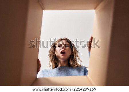 Beautiful woman opening cardboard box in shock face, looking skeptical and sarcastic, surprised with open mouth 