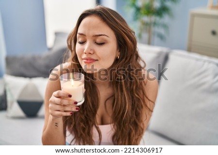 Young beautiful hispanic woman sitting on sofa smelling aromatic candle at home Royalty-Free Stock Photo #2214306917