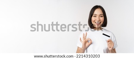 Smiling korean girl showing okay sign and recommending credit card of copy space bank, standing in tshirt over white background
