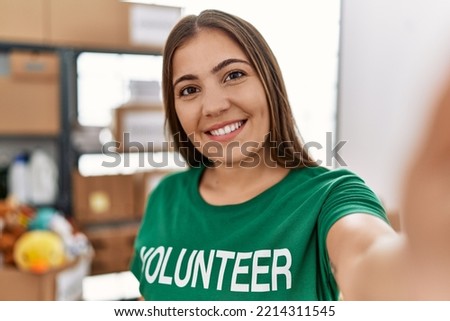 Young hispanic woman wearing volunteer uniform making selfie by the camera at charity center