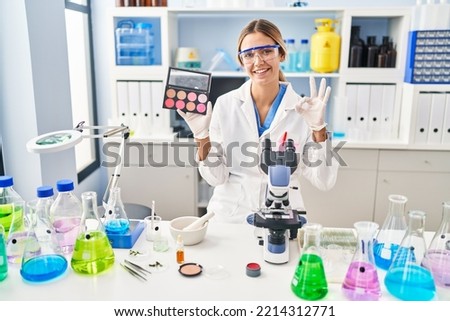 Young blonde woman working at scientist laboratory with make up doing ok sign with fingers, smiling friendly gesturing excellent symbol 