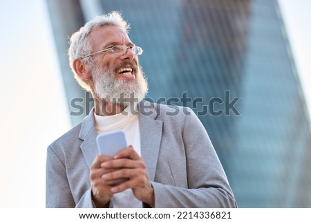 Happy older mature stylish professional business man, smiling senior 60 years old businessman wearing suit holding smartphone using mobile cell phone tech standing outdoor in big city office district. Royalty-Free Stock Photo #2214336821