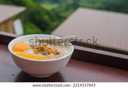 Horizontal picture top view, pork porridge with eggs in a white plate for breakfast, placed on a brown table. There is a blurred background of a view homestay,  in holiday