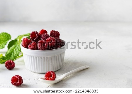 Ripe juicy raspberries in a white ceramic vase on a light table. Green leaves in the background. A white spoon on the table. Light background, space for text