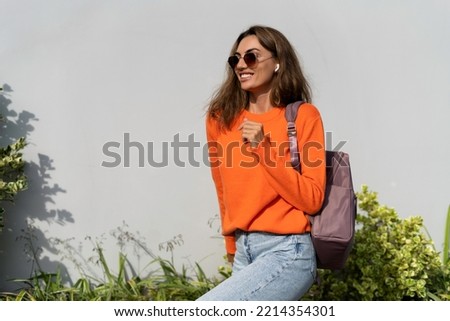 Woman in casual orange sweater and sunglasses on gray background, wearing headphones listening to music dancing cheerful happy ad shocked surprise excited copy space purple backpack on her shoulder