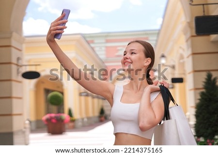 happy girl, young beautiful woman is smiling taking selfie picture of herself on smartphone camera, using cell mobile phone in mall during shopping holding shopping paper bags in hand outdoors.