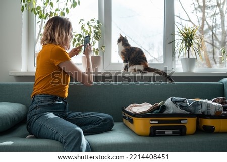 Young woman sitting on sofa taking photo of cat on window sill. Packing the baggage. Preparing for trip, journey, travel.
