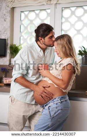 Beautiful young couple with pregnant wife standing in the kitchen with her husband touching his wife's bump and kissing her in the forehead while she holds a coffee cup enjoying the moment.