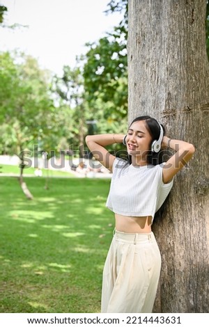 Portrait, Calm and relaxed young Asian female listening to music through her headphones, leaning on the tree, eyes closed, chilling in the green park.