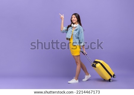 Full body portrait of young pretty Asian woman tourist with suitcase pointing finger up in purple color studio isolated background