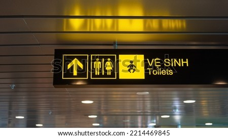 Toilets with arrow and gender icon  lighting signboard (English and Vietnamese text) which is installed on ceiling of the airport terminal building, selective focus.